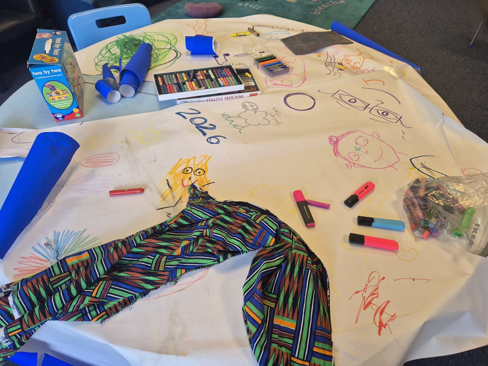 A table covered with children's drawings, oil pastels, highlighters and colourful fabric during a creative session